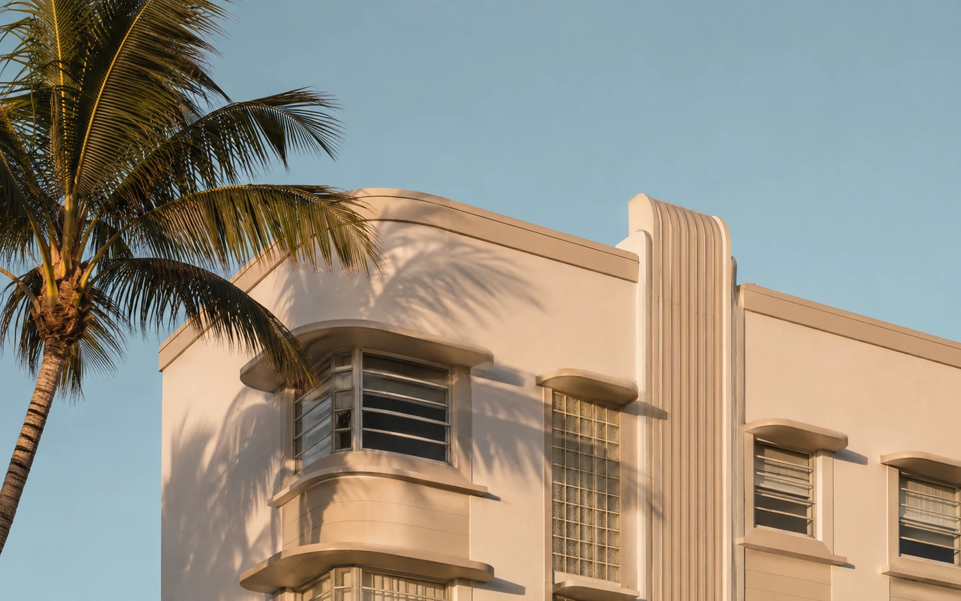 Restored Art Deco facade in Miami Beach in late afternoon light, with a curved corner, eyebrow window shades and glass block detail behind a mature palm