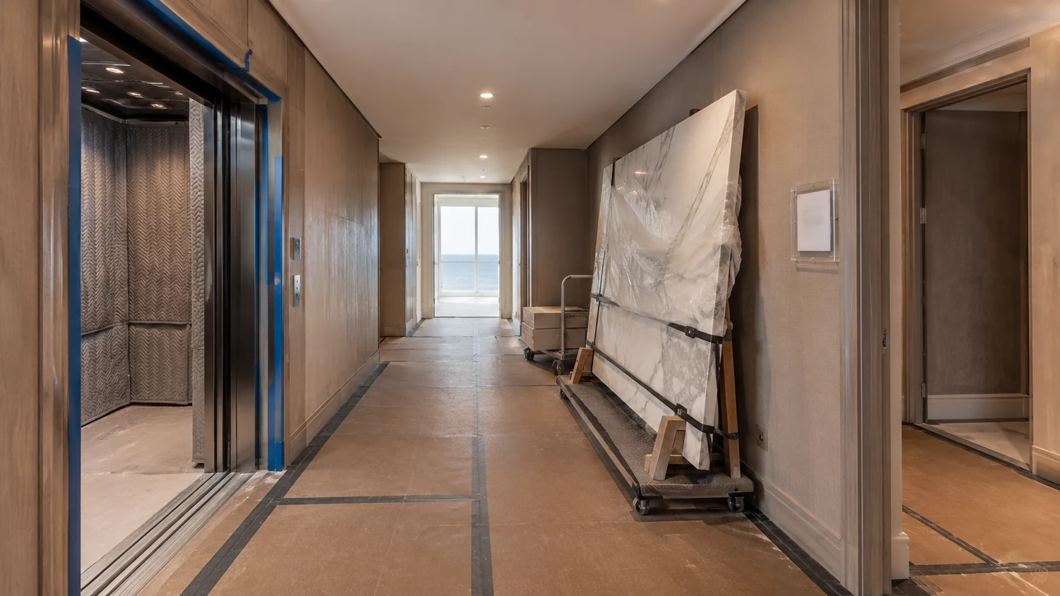 Service corridor of a Miami high rise condominium during a renovation, with the freight elevator open and lined with quilted protection pads, brown floor board taped over the floor, and a film wrapped stone slab strapped upright to a trolley beside the wall
