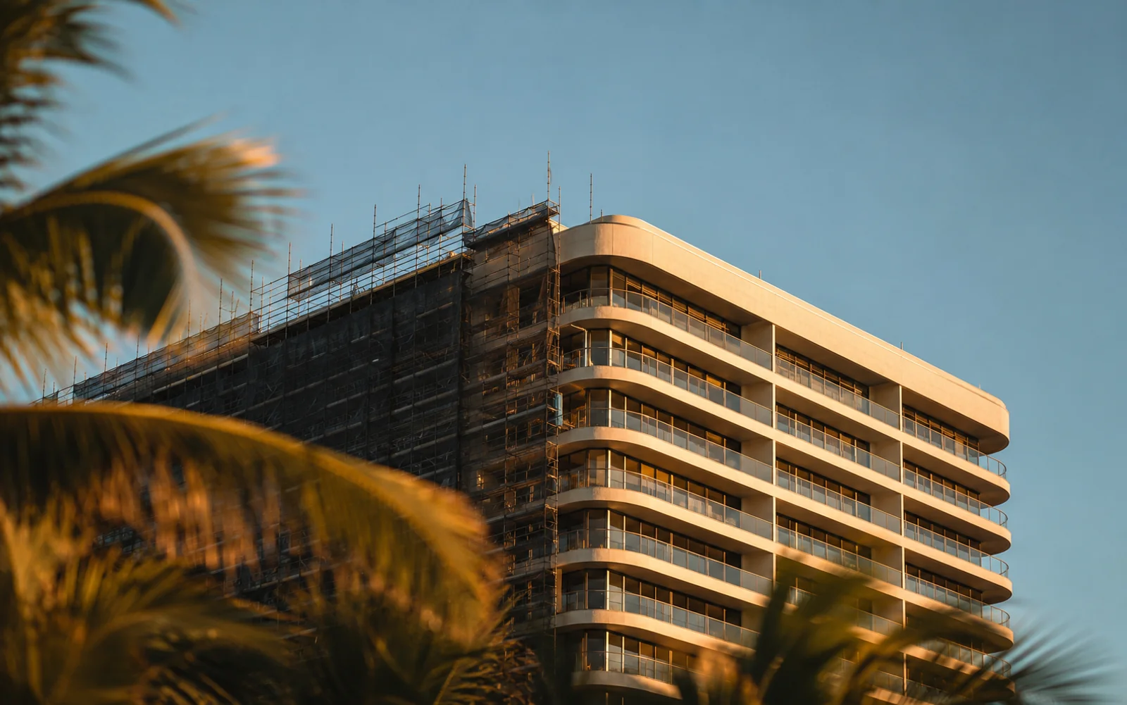 Oceanfront residential tower in Miami at golden hour, half covered in construction scaffolding and half showing restored concrete and new glass balconies during a building recertification cycle