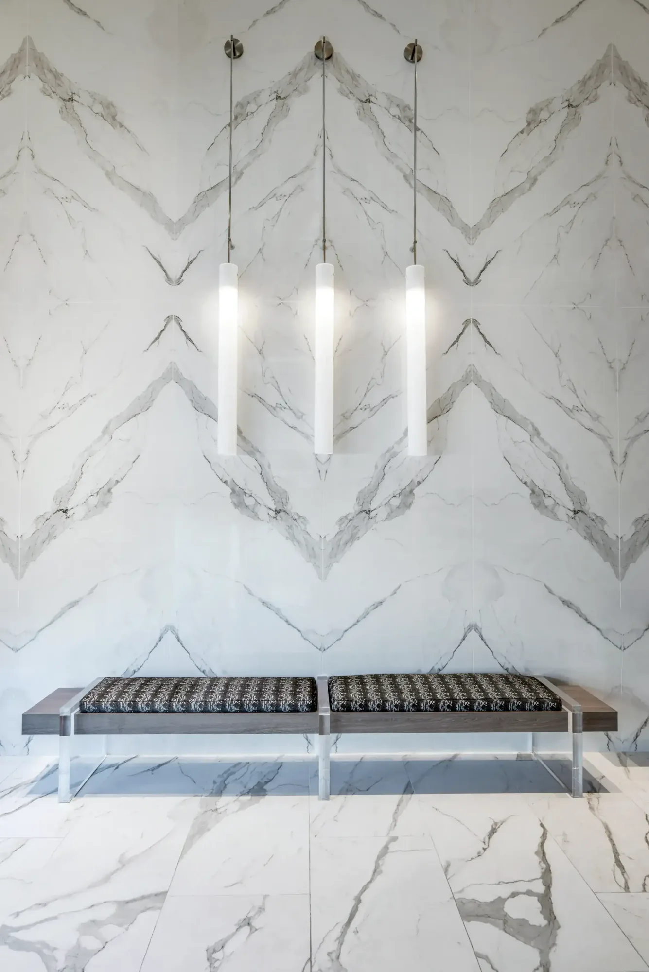 Upholstered bench against a chevron marble wall at the Diplomat Residences in Hollywood, Florida, under three cylindrical pendants