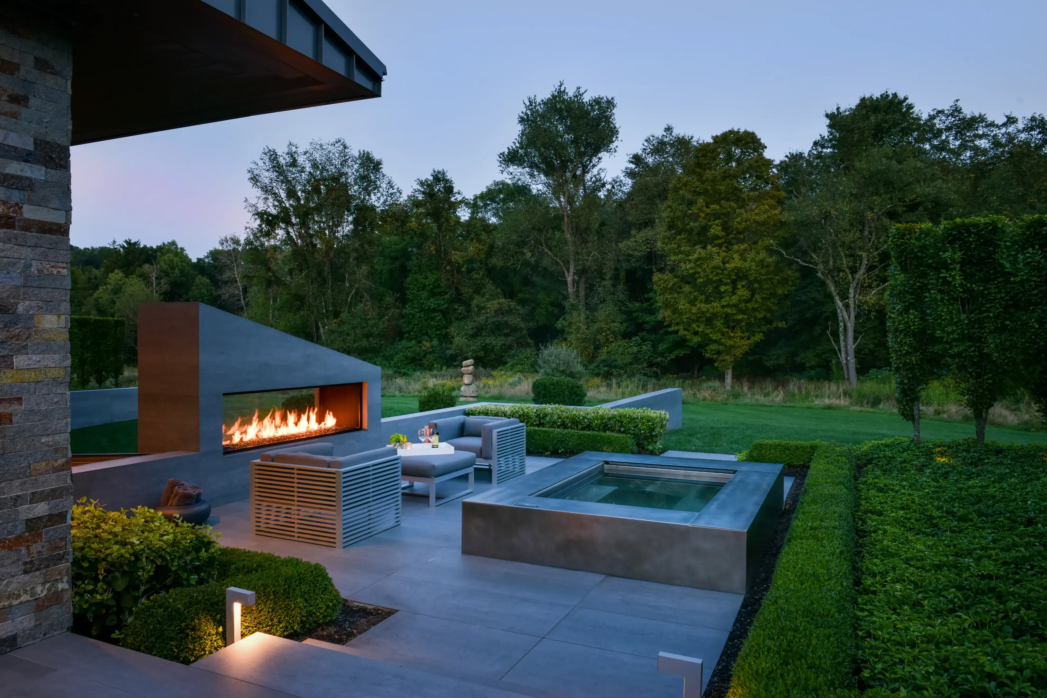 Outdoor terrace at the Richfield Residence in Davie, Florida, at dusk, with a linear fire feature, spa and clipped hedging